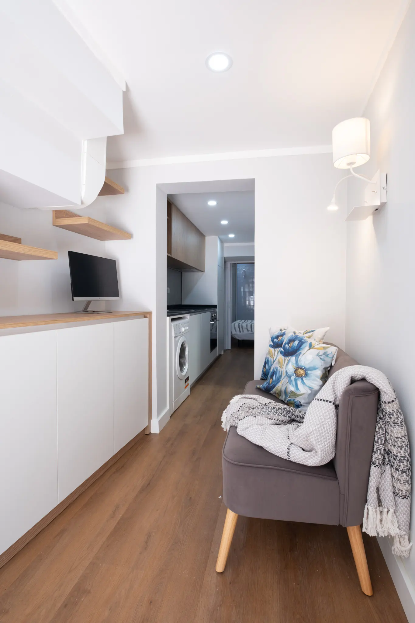 A modern studio apartment with wood floors, a gray sofa with floral pillows, and a white kitchen in the background.