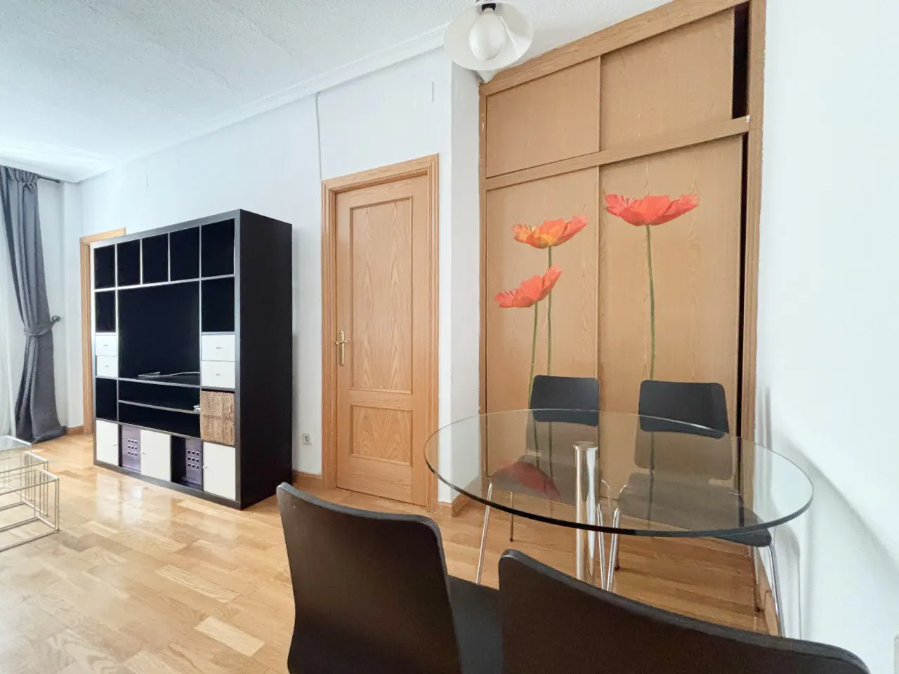 Dining area with a glass table, four black chairs, a black shelving unit, and wooden doors with poppy flower decals.