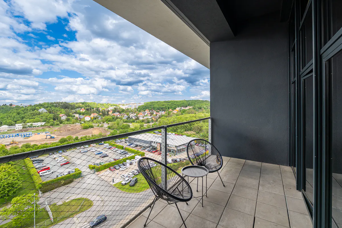 Balcony view with two black chairs and a small table overlooking a parking lot, trees, and a cloudy sky.