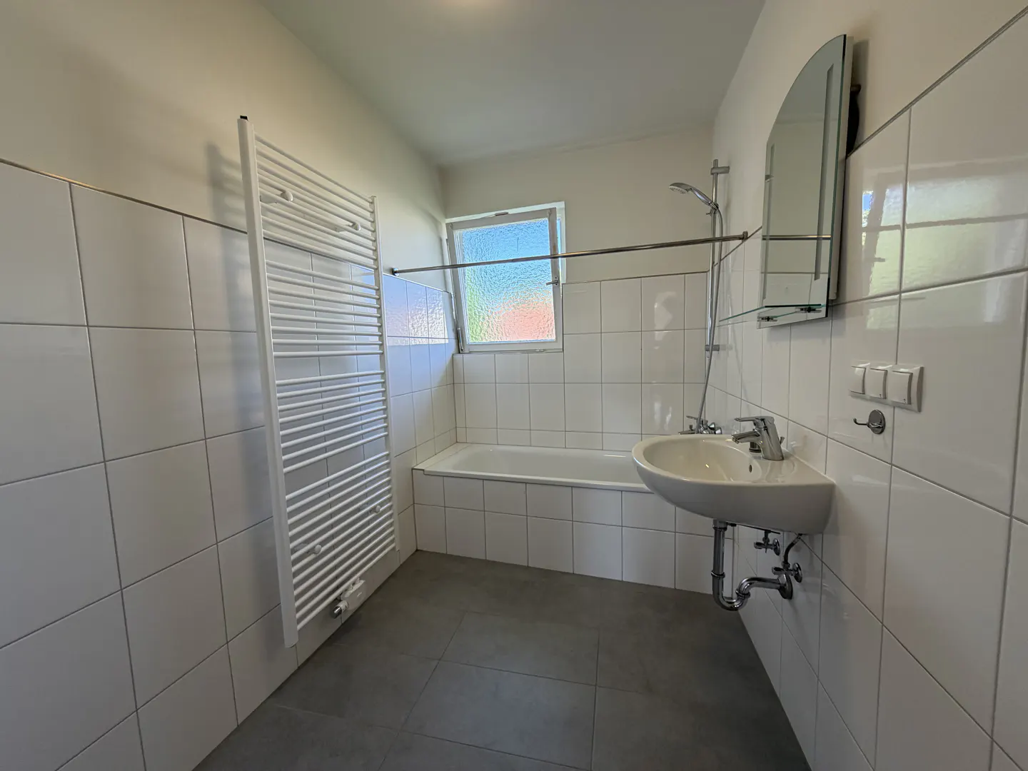 Bathroom with white tiled walls, grey floor, bathtub, wall-mounted sink, and a tall white towel radiator.