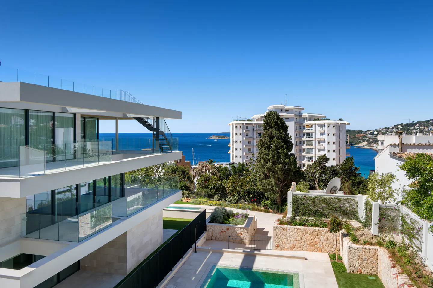 Modern white villa with glass balconies overlooks a turquoise pool and the blue sea under a clear sky.