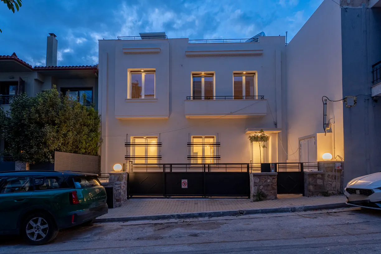 Two-story white house with black gate at dusk. Warm light shines from the windows. Cars are parked on the street.