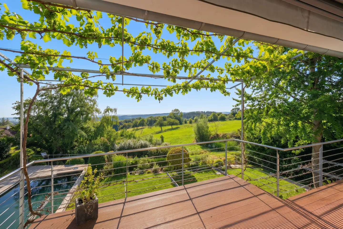 A wood deck with a metal railing overlooks a green landscape. A vine-covered trellis provides shade.
