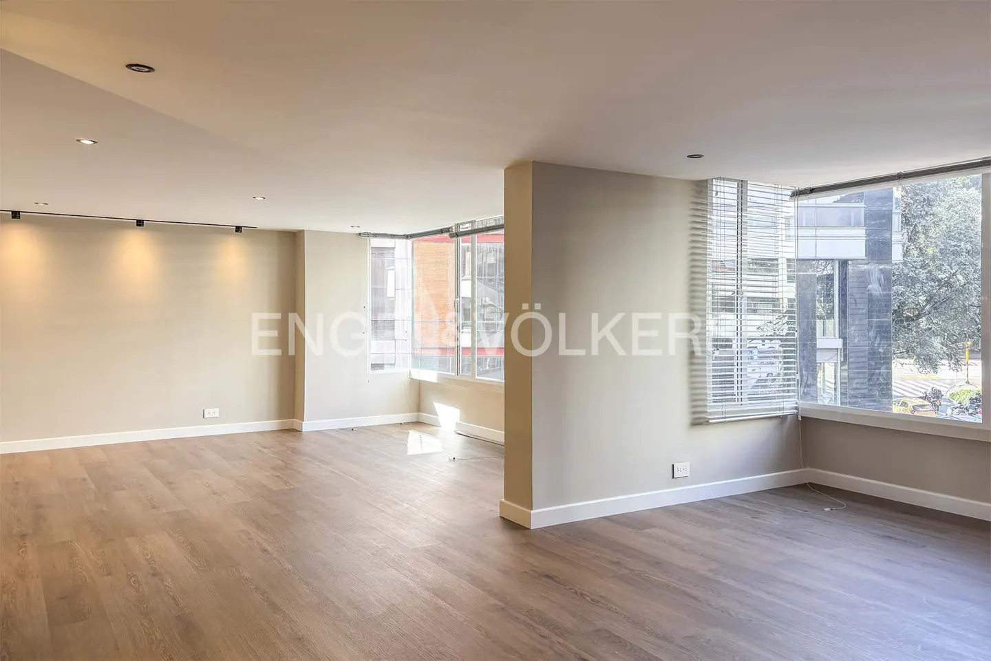 Empty, light-filled room with wood floors, beige walls, and large windows with blinds. Track lighting on the ceiling. Engel & Völkers logo visible.