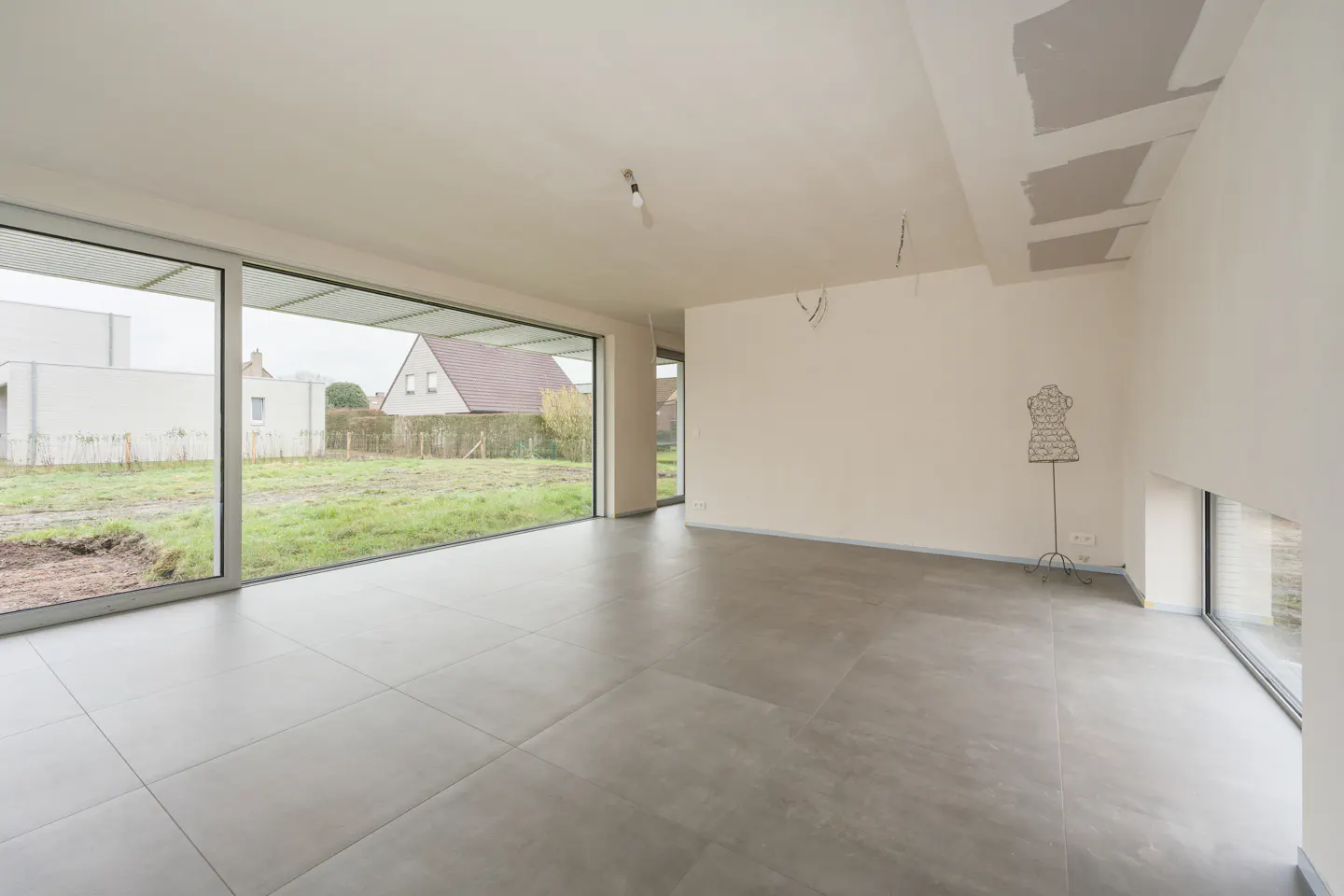 Unfurnished room with gray tile floor, white walls, and large windows overlooking a yard with grass and houses. A dress form stands in the corner.