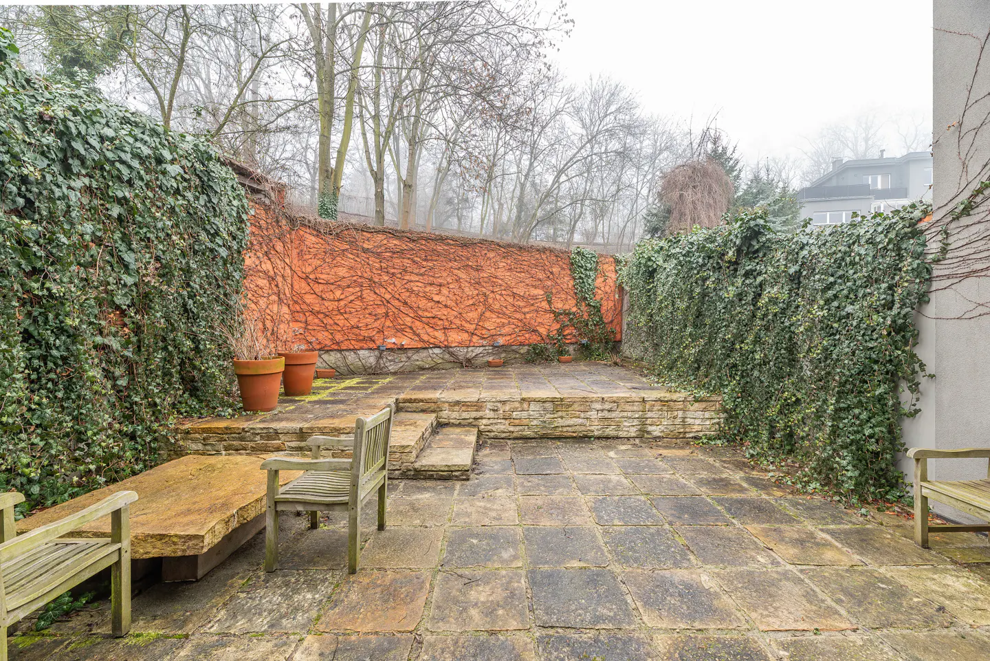 Stone patio with wooden benches, surrounded by ivy-covered walls and trees in the background. The sky is overcast.