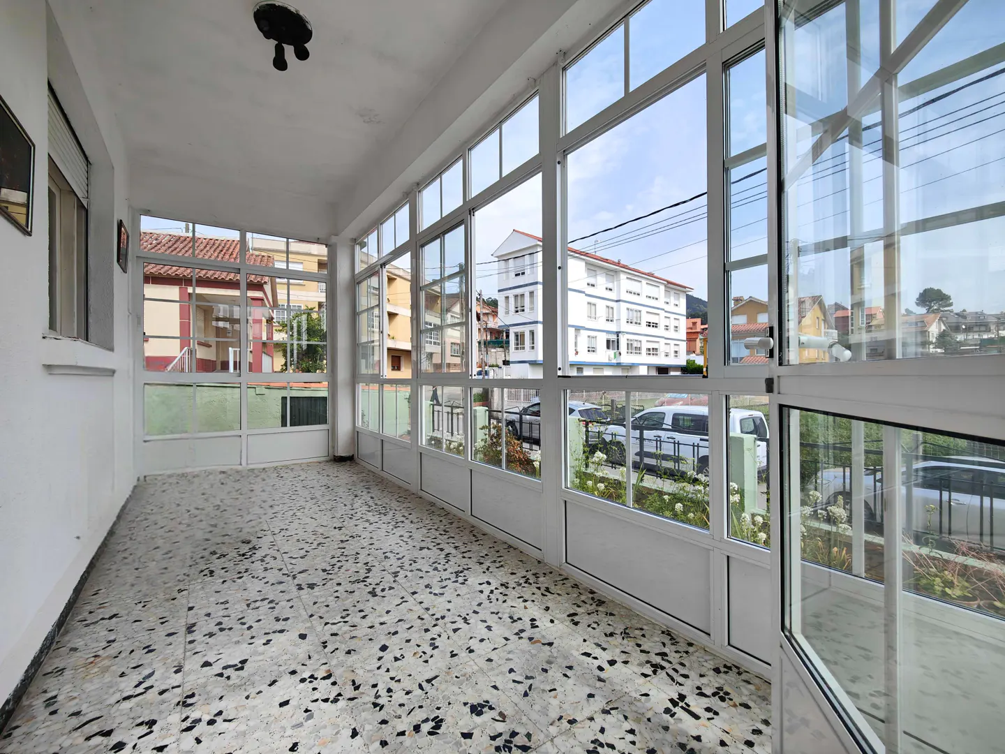 Sunroom with white walls, terrazzo floor, and large windows showing a street with houses and cars.