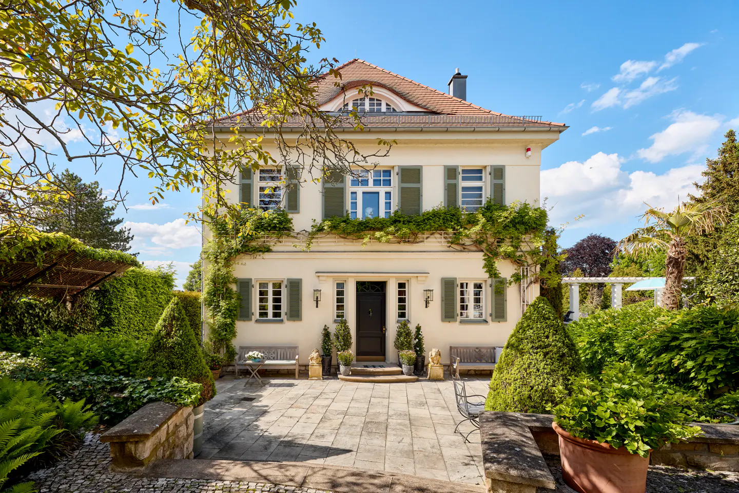 Cream-colored two-story villa with green shutters, a tiled roof, and a stone patio surrounded by lush garden greenery.