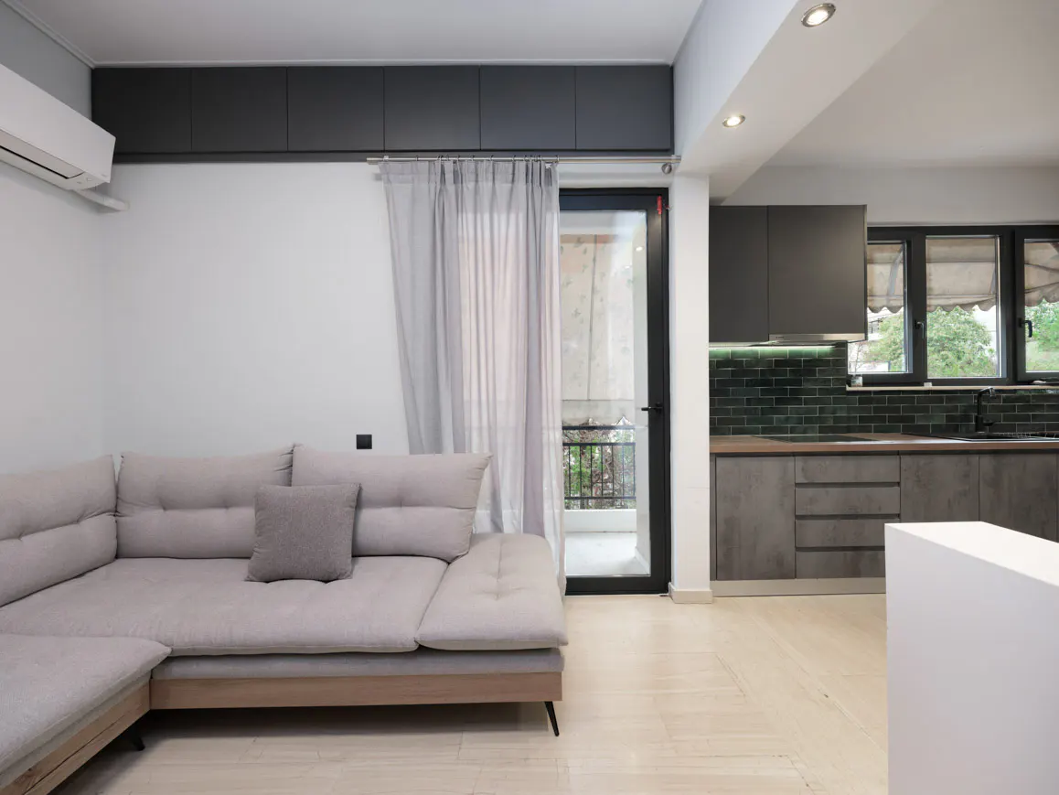 Modern apartment interior with a gray sectional sofa, balcony door with sheer curtains, and a gray kitchen with dark green backsplash.