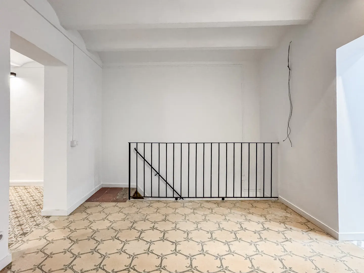 Bright room with white walls, patterned tile floor, and black metal railing. An open doorway leads to another room.