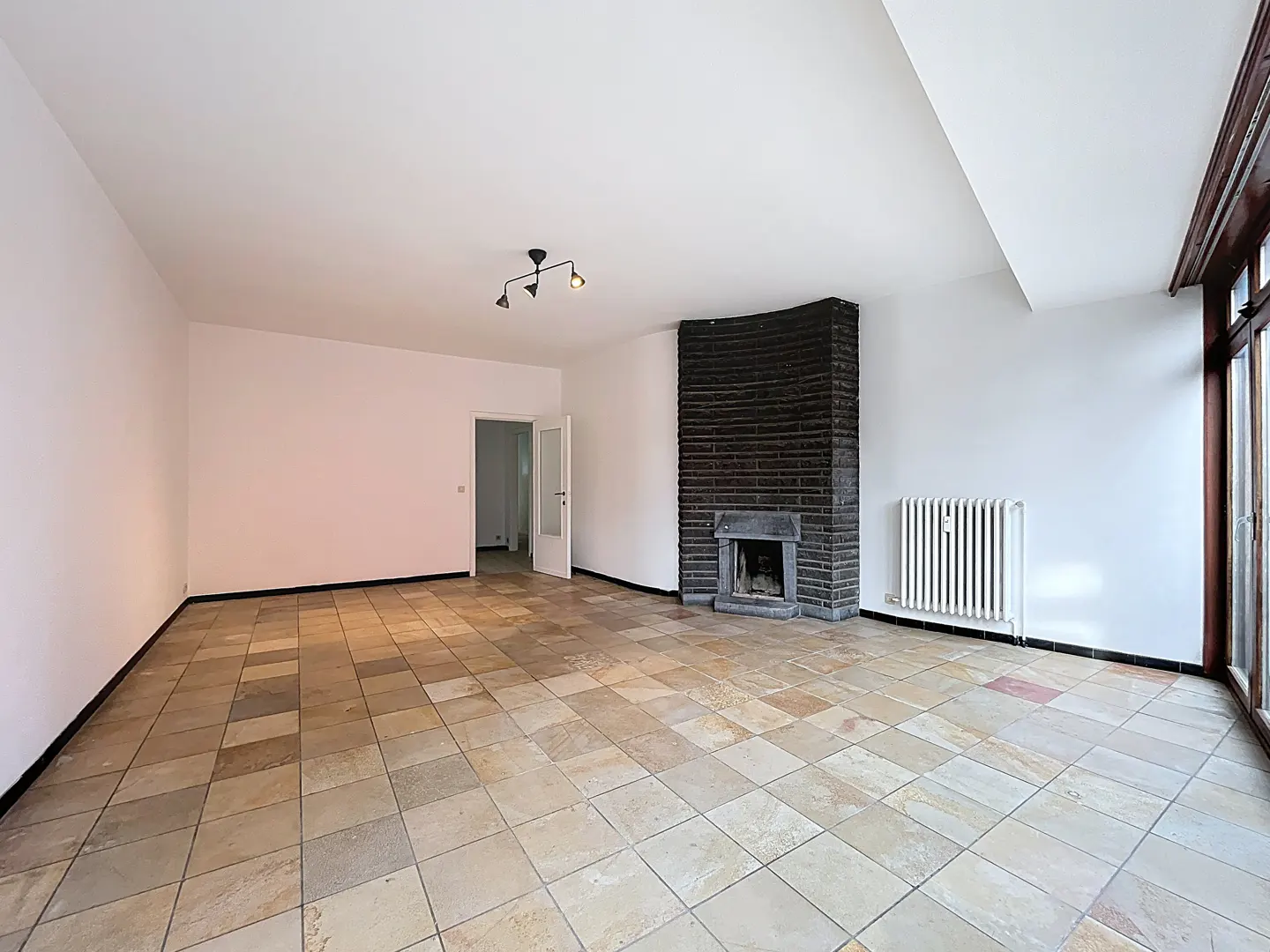 Empty room with tan tile floor, white walls, black brick fireplace, radiator, and light fixture.