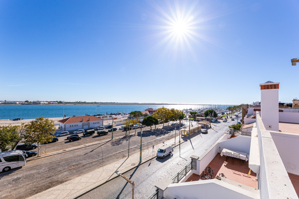 High-angle view of a sunny coastal street with parked cars, trees, and a marina under a bright blue sky. [AI-generated text]