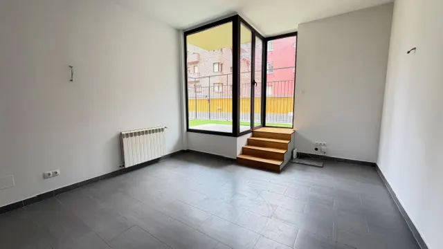 Bright, empty room with gray tile floor, white walls, radiator, and three wooden steps leading to a window with a view of a courtyard.