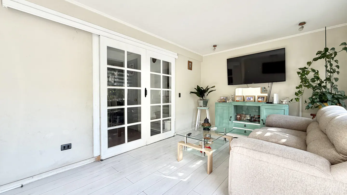Living room with white wood floors, a beige sofa, glass coffee table, and a light green cabinet under a wall-mounted TV. White sliding glass doors are on the left.