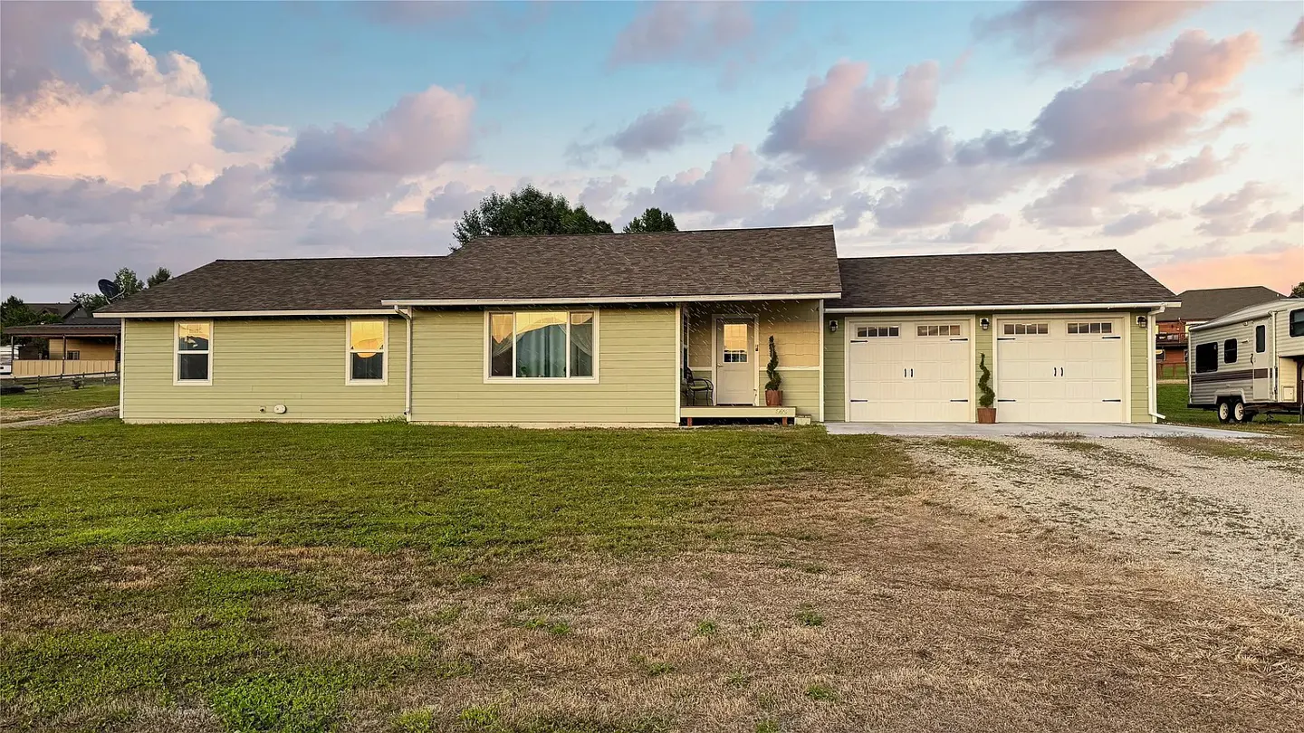 A single-story, light green house with a brown roof, a two-car garage, and a gravel driveway. A camper is parked to the right.
