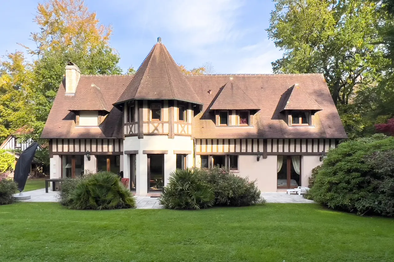 Exterior view of a two-story Tudor-style house with a brown roof, beige walls, and dark wood trim, surrounded by green lawn and trees.