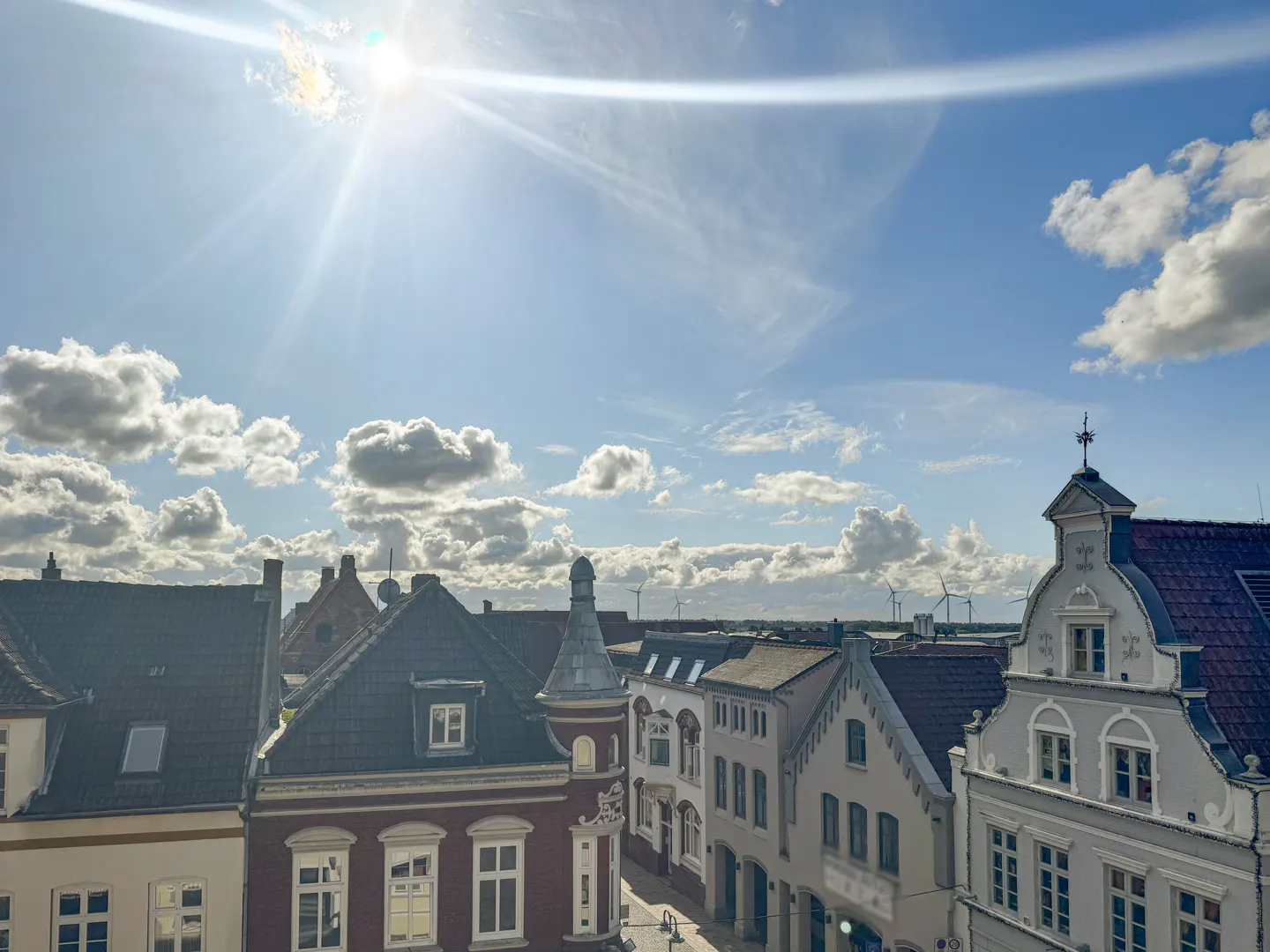 Rooftop view of a European town under a bright, sunny sky with scattered clouds. Wind turbines are visible in the distance.