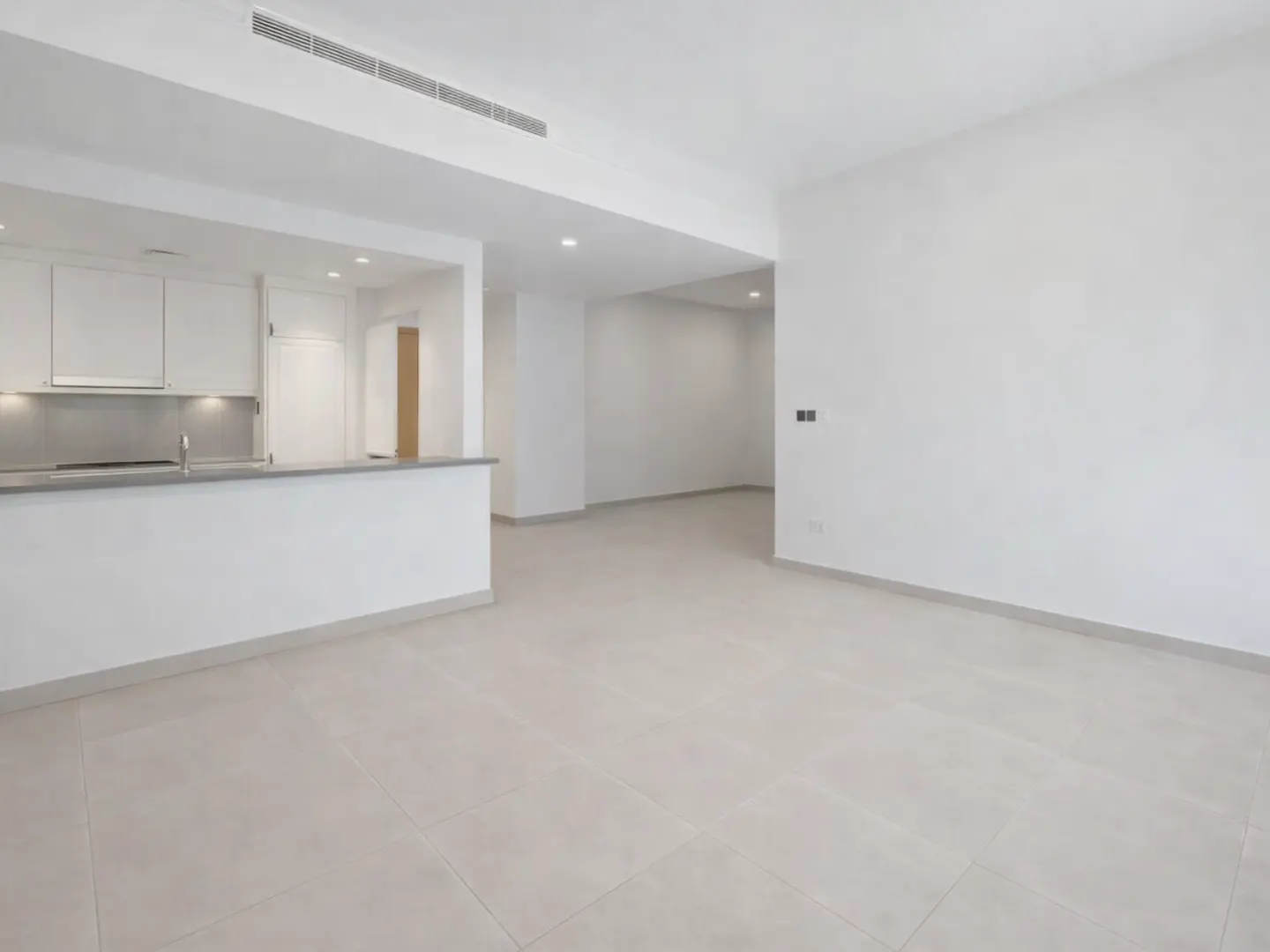 A bright, empty apartment with white walls and tile floors. An open kitchen with white cabinets is visible in the background.