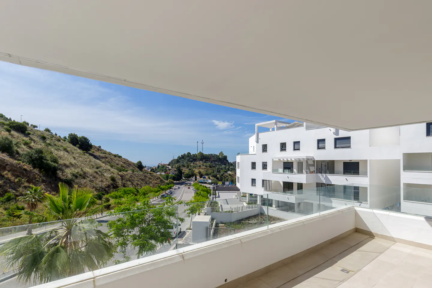 View from a balcony with glass railings, overlooking a white building, a road, and a green hillside under a blue sky.