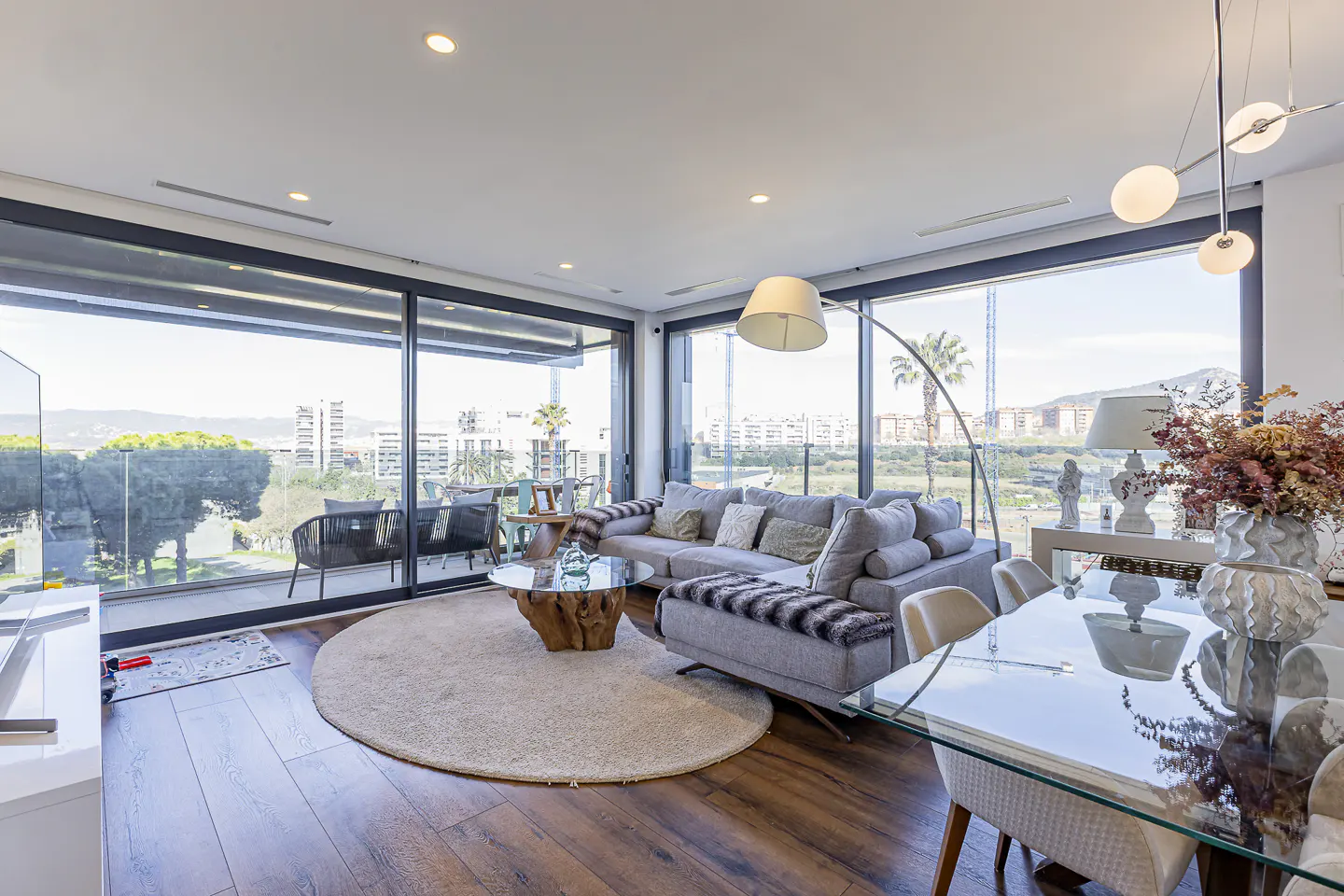 Bright living room with wood floors, a gray sofa, and a round rug. Large windows offer city views. A glass table and chairs are in the foreground.