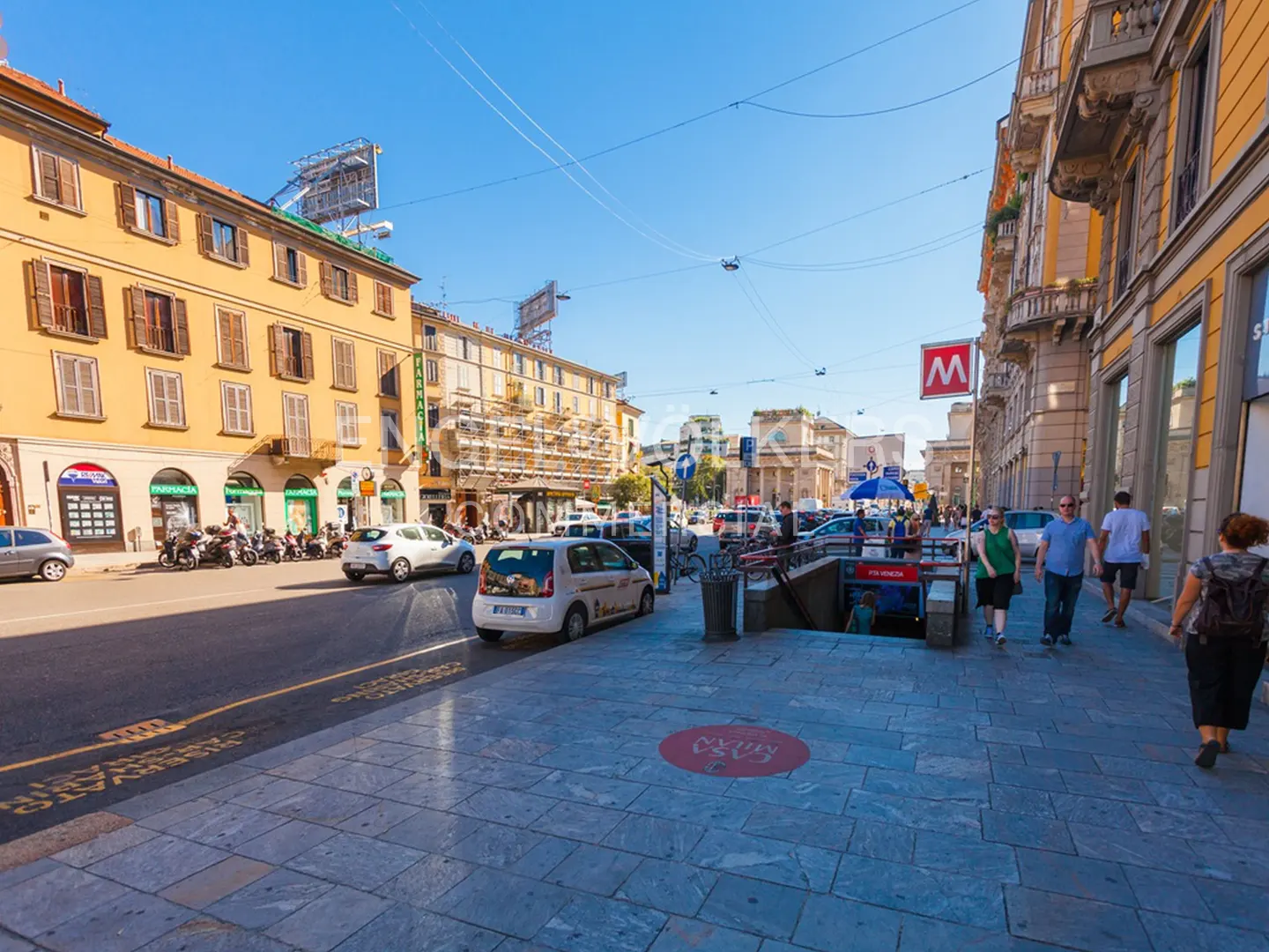 Street view in Milan, Italy, with yellow buildings, cars, and people walking near a metro station entrance.