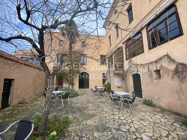 Outdoor courtyard with stone paving, tables, and chairs. A bare tree stands in the foreground, with a tan building in the background.