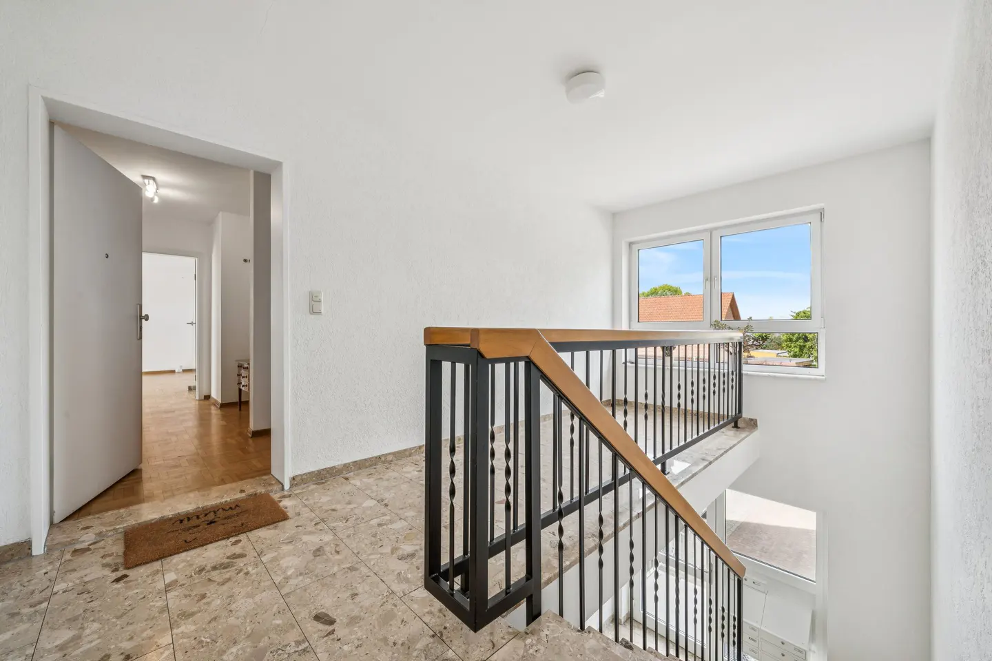 Bright hallway with stone tile floors, a wooden-railed staircase, and a doorway leading to a room with wood flooring.
