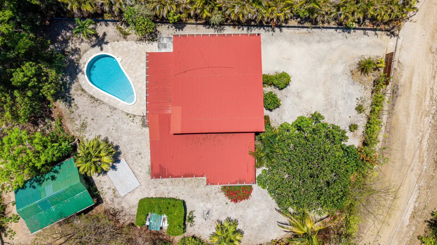 Aerial view of a red-roofed house with a blue pool, surrounded by green trees and sandy ground.