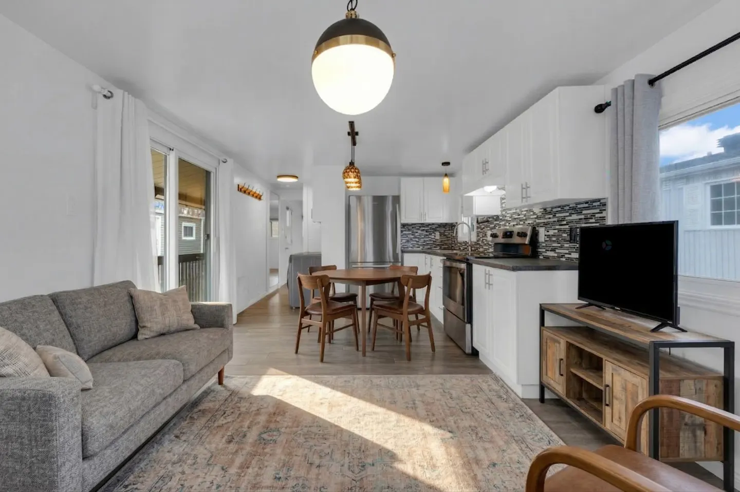 Bright, open-concept living space with gray sofa, wood table, white kitchen cabinets, and a TV on a rustic stand.