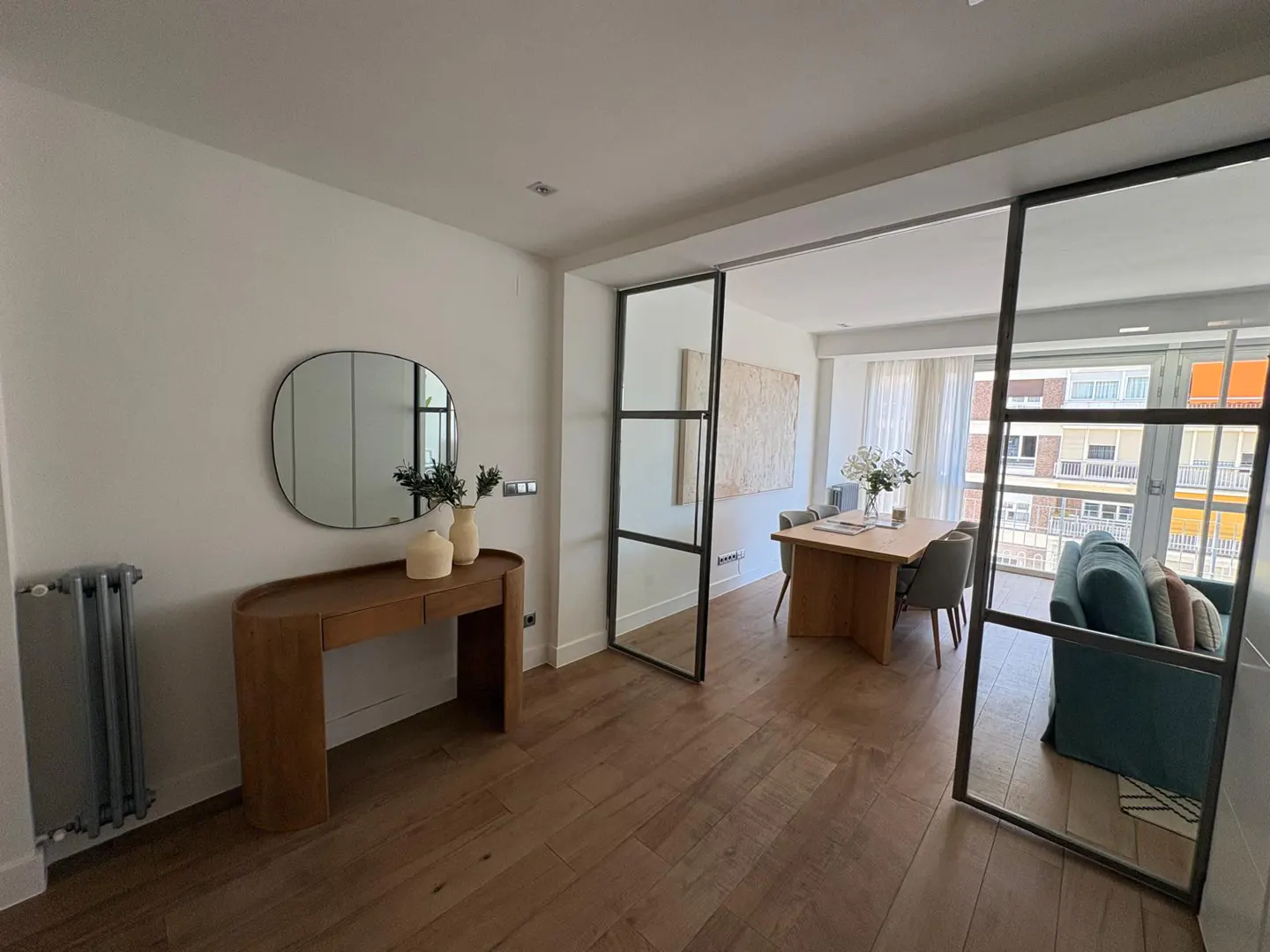 Modern apartment with a wooden console, oval mirror, and a glass-partitioned dining area with a blue sofa and balcony view.