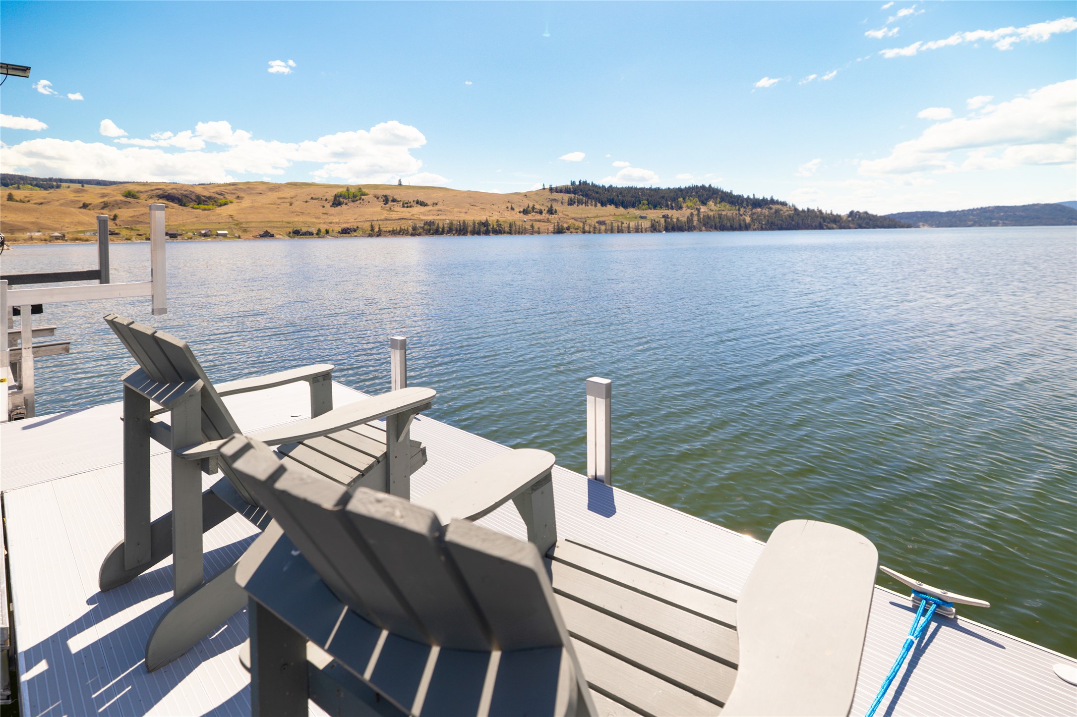 Two gray Adirondack chairs on a dock overlook a lake with rolling hills under a blue sky.