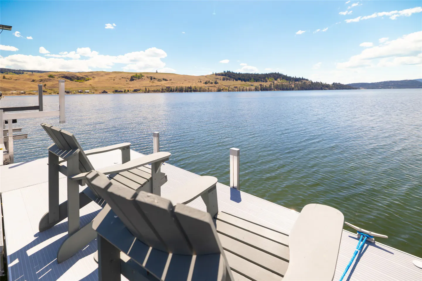 Two gray Adirondack chairs on a dock overlook a lake with rolling hills under a blue sky.