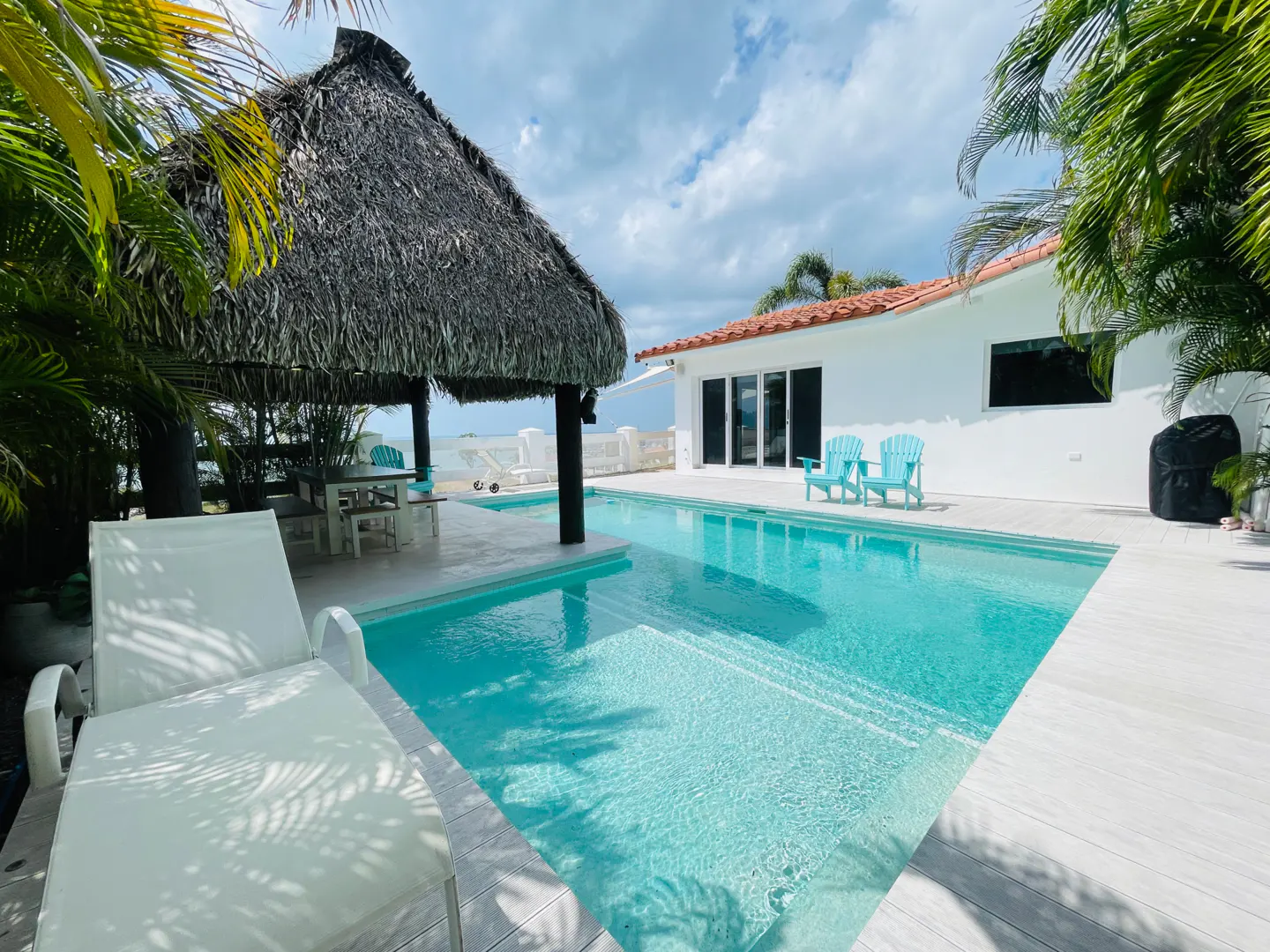 Outdoor pool with a thatched-roof cabana, white lounge chairs, and a white house with a red tile roof.