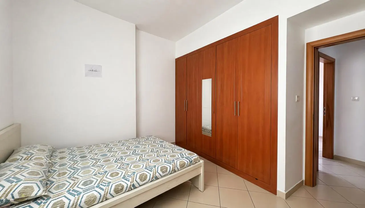 Bedroom with a white-framed bed, patterned bedding, and a large wooden wardrobe with a mirror. A doorway leads to another room.