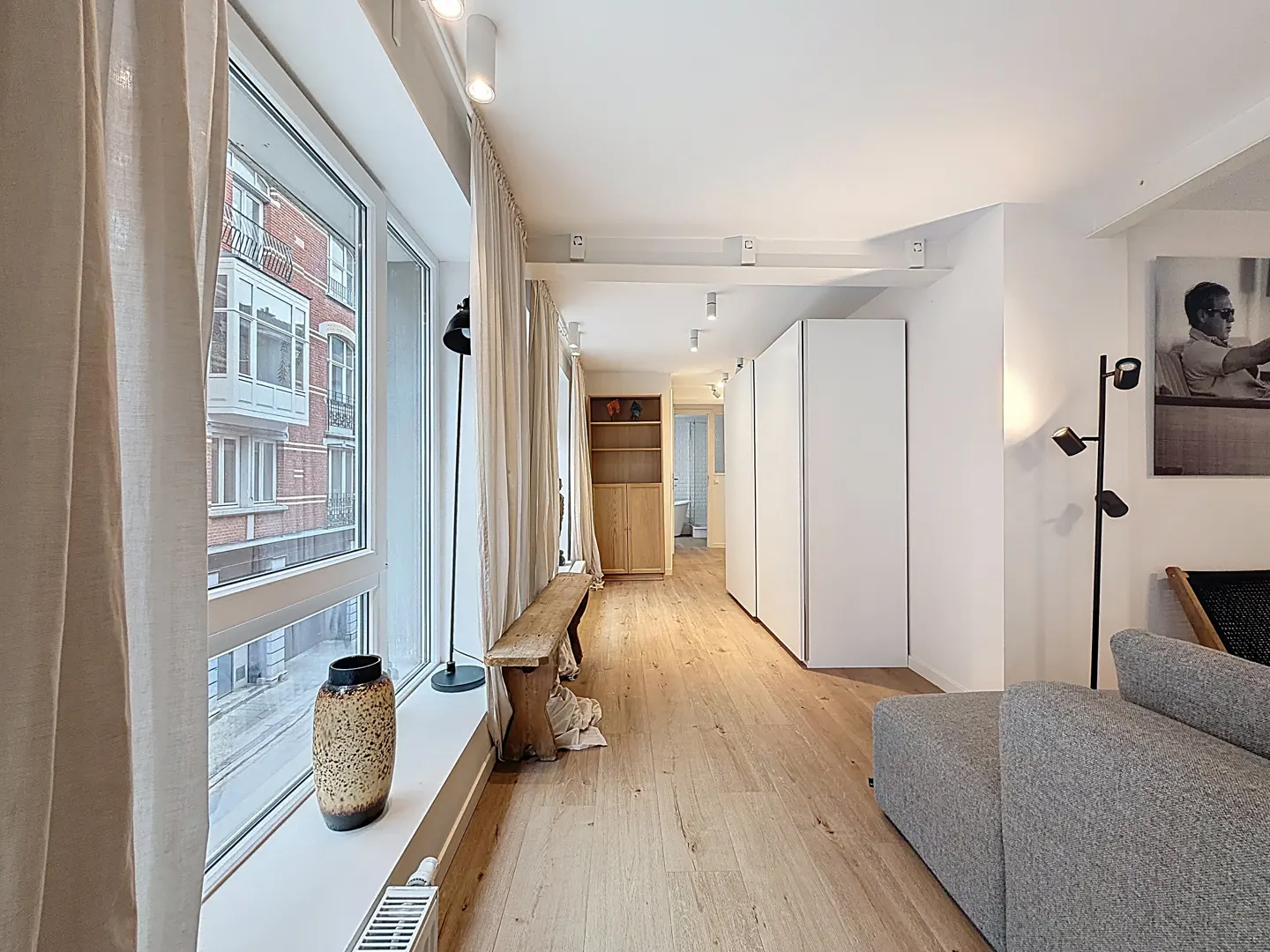 Bright, modern apartment interior with wood floors, white walls, and large windows. A gray sofa and wooden bench add warmth.