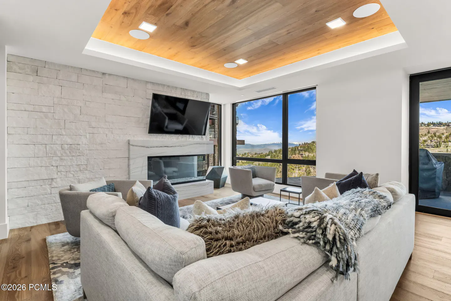 Living room with a gray sofa, stone fireplace, and large windows with a mountain view. The ceiling has wood paneling and recessed lighting.