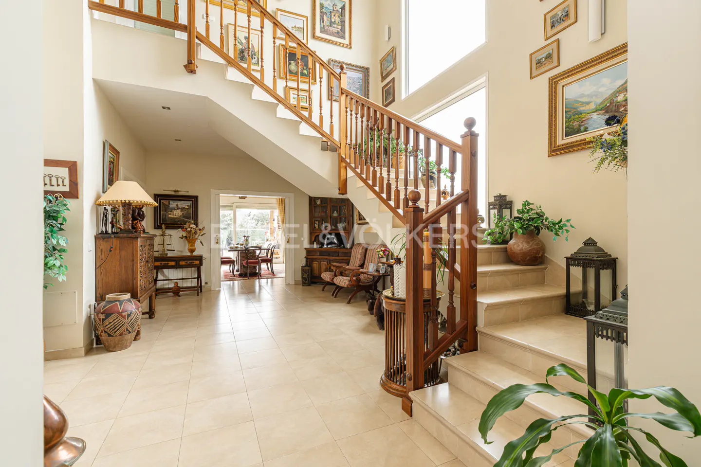 Grand foyer with a staircase, wooden banister, and art-lined walls. Beige tile flooring and a glimpse of the dining room.