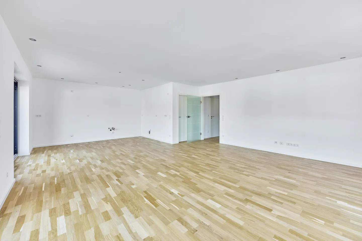Bright, empty room with light wood floors and white walls. Glass doors lead to other rooms.