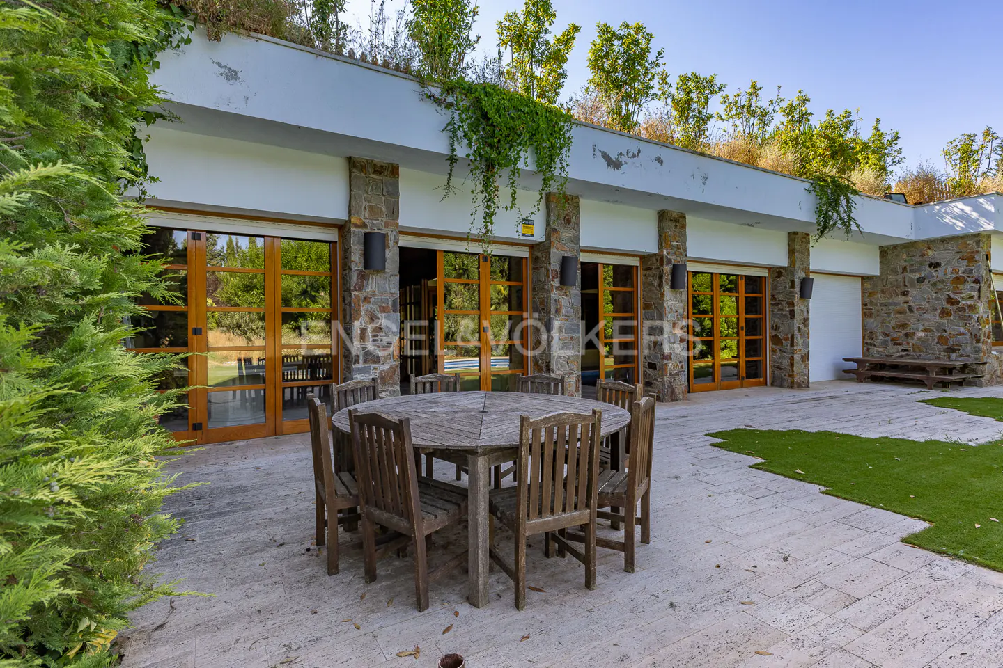 Outdoor patio with a round wooden table and chairs, stone pillars, and large glass doors. Greenery grows on the roof.