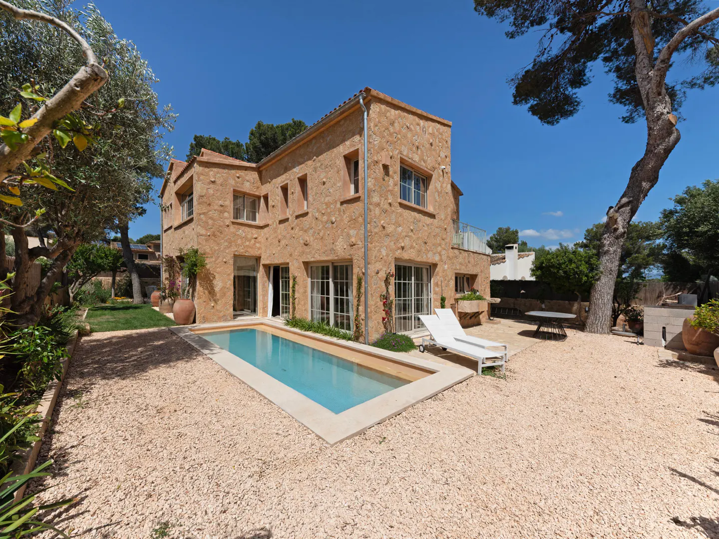 Exterior view of a two-story stone house with a rectangular pool, white lounge chairs, and a gravel yard under a blue sky.