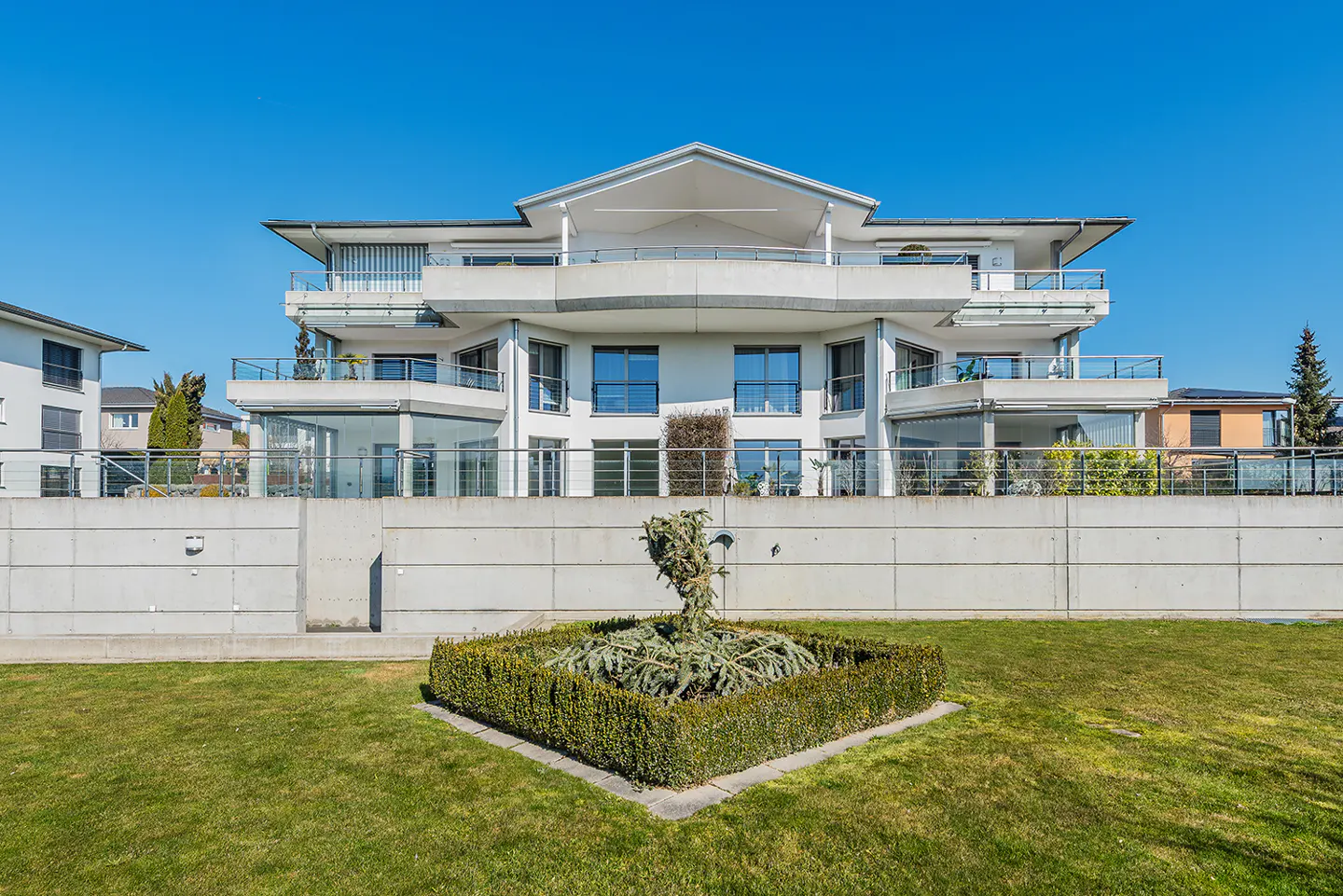 Three-story white building with balconies, behind a concrete wall and green lawn. Blue sky.