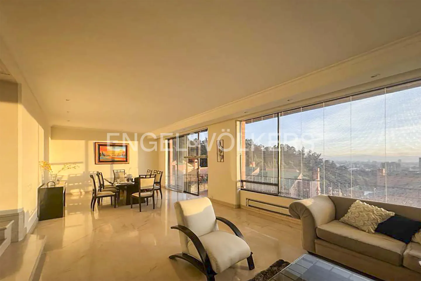 Spacious living and dining room with cream furniture, marble floors, and large windows overlooking a city view at sunset.