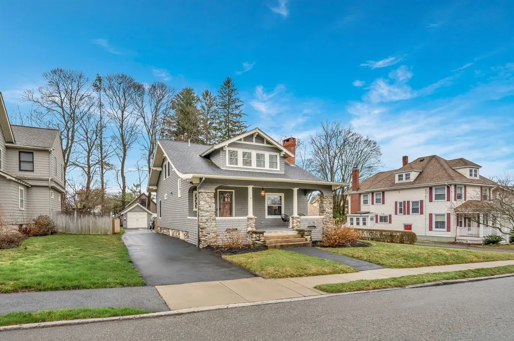Gray two-story house with stone accents, a front porch, and a driveway leading to a detached garage. Blue sky background.