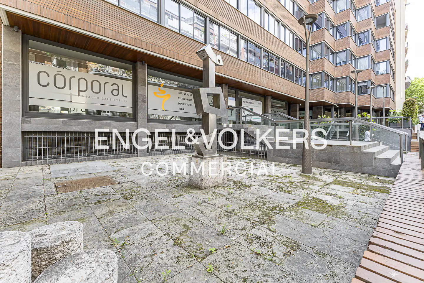 Exterior of a brick building with "Engel & Völkers Commercial" text overlay. A modern sculpture stands in front.