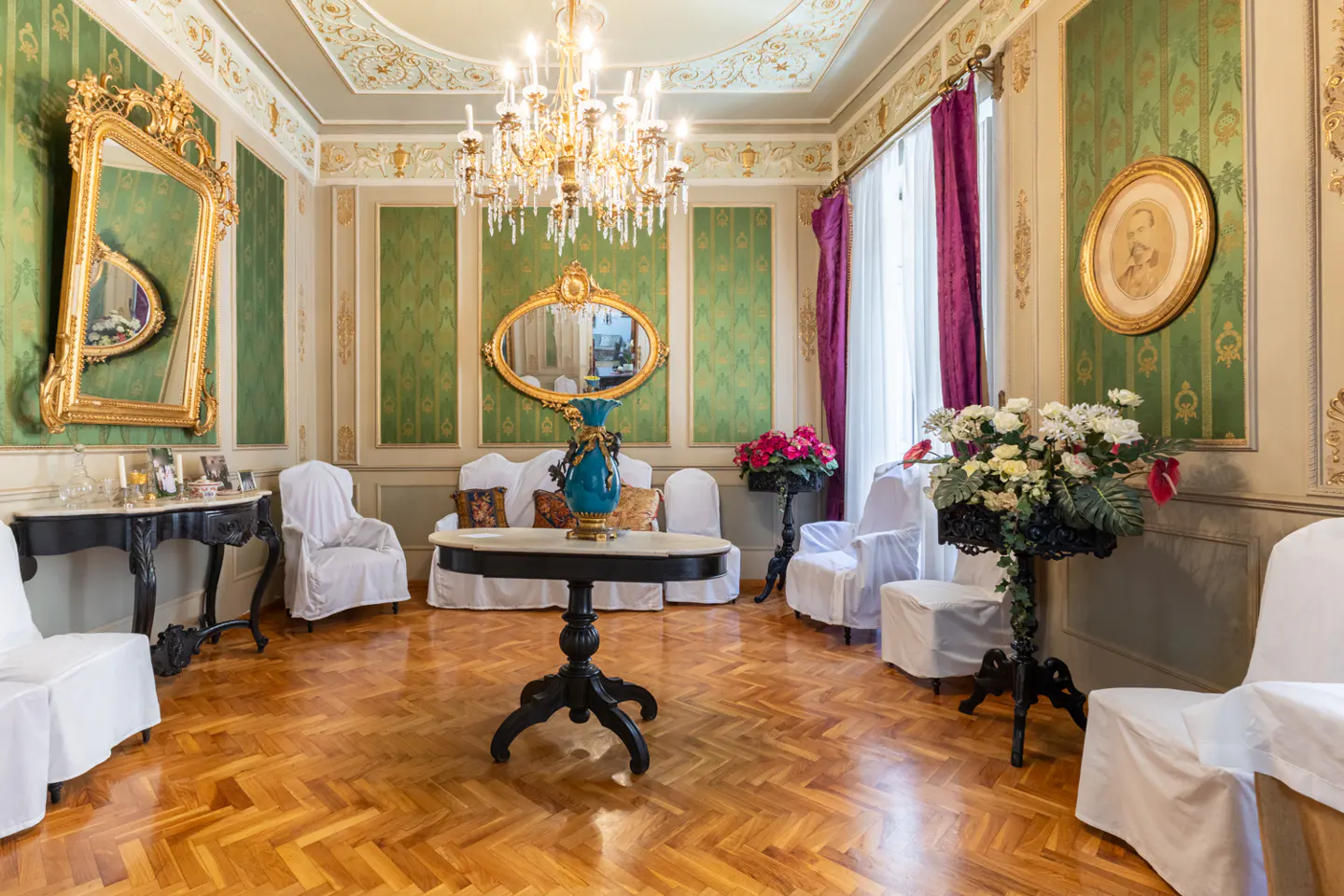 Ornate room with green walls, gold trim, and herringbone wood floors. White furniture, mirrors, chandelier, and purple curtains add elegance.