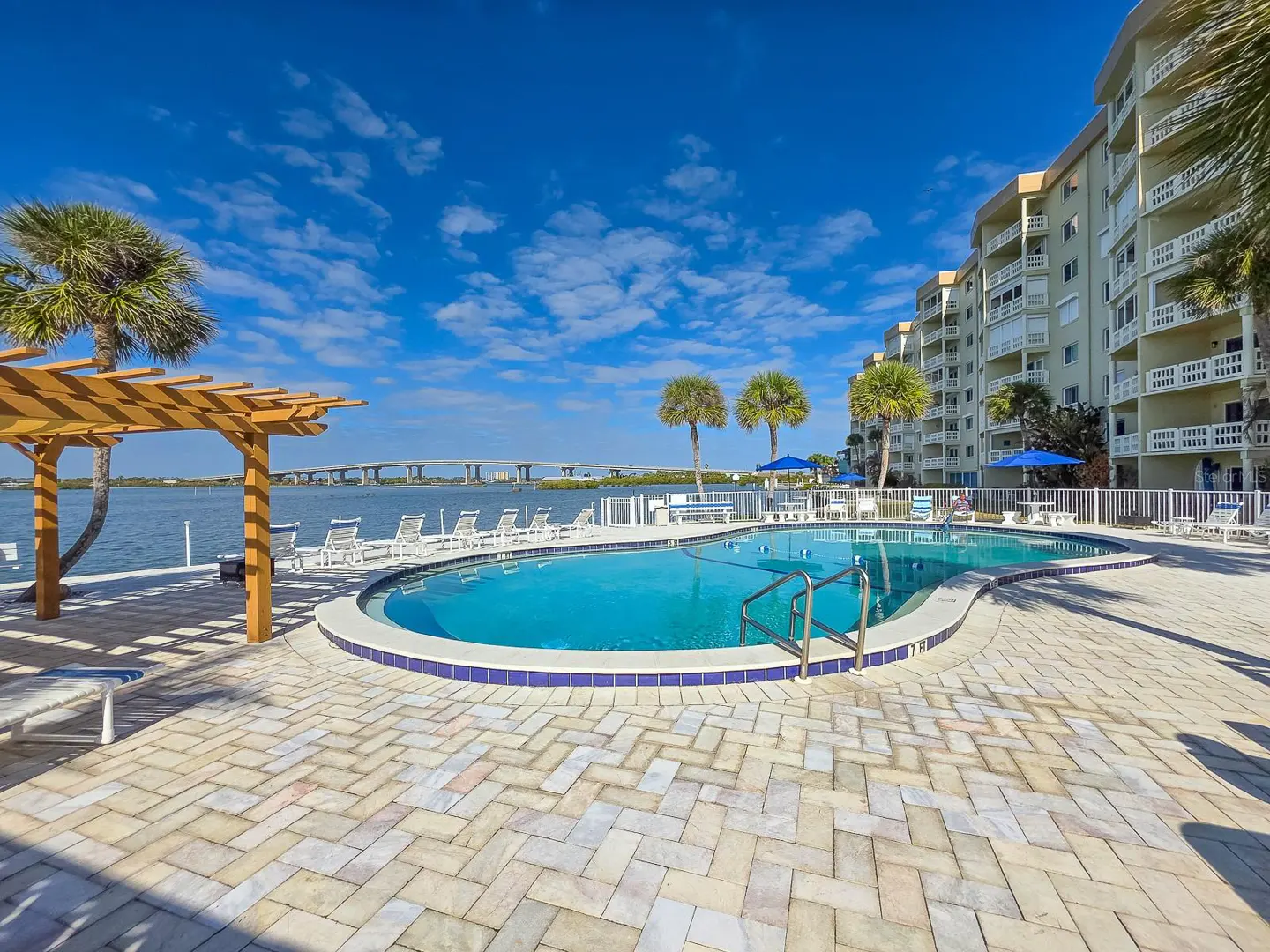 Outdoor pool with blue water, surrounded by palm trees, lounge chairs, and a building with balconies under a bright blue sky.