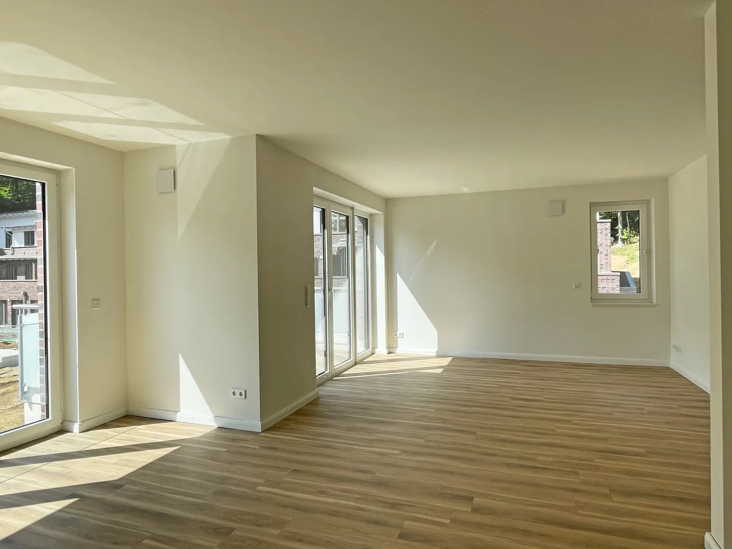 Bright, empty room with light wood floors, white walls, and sunlight streaming in through windows and sliding glass doors.