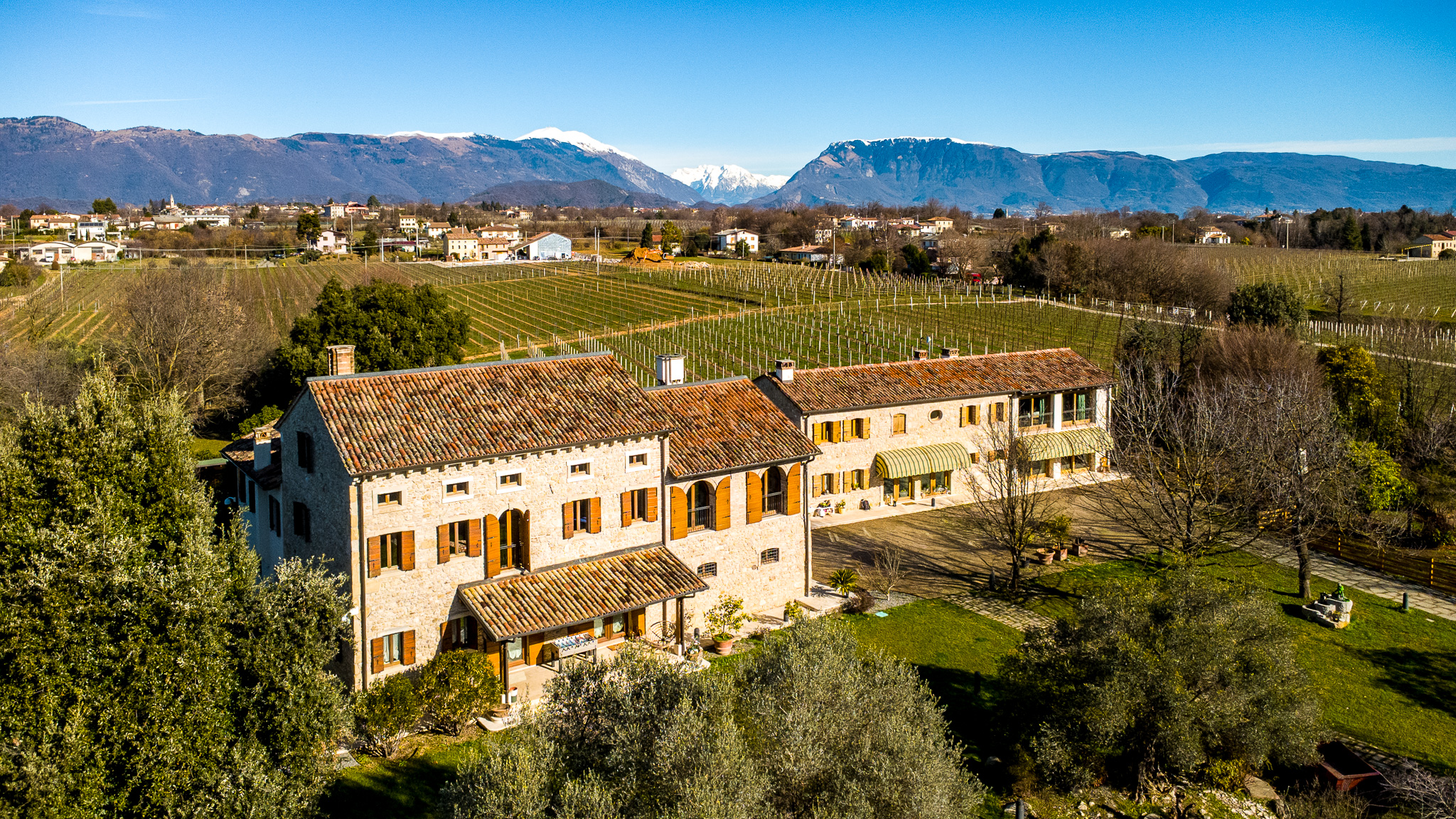 Aerial view of a stone house with orange shutters, surrounded by vineyards and mountains under a blue sky.