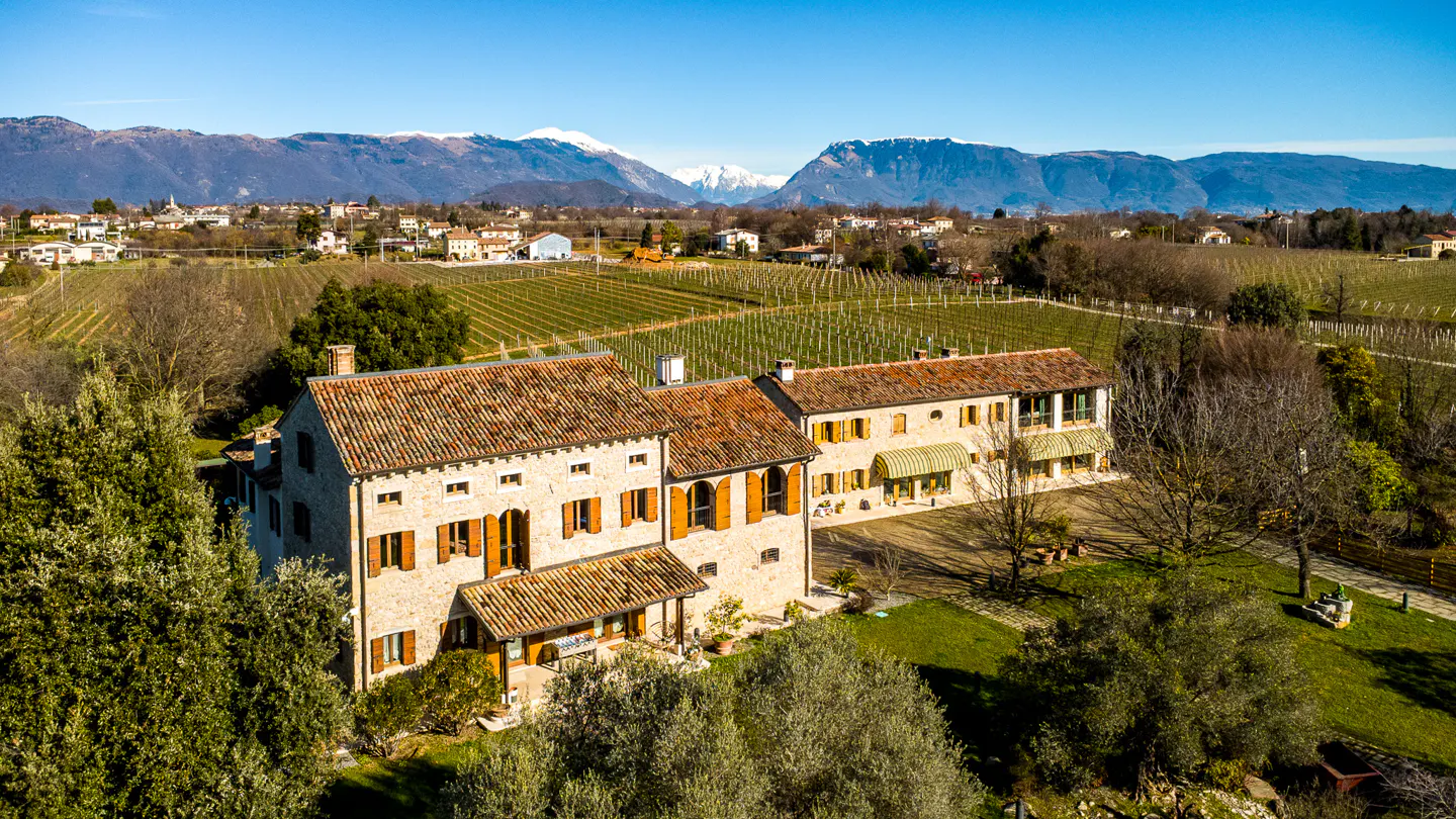 Aerial view of a stone house with orange shutters, surrounded by vineyards and mountains under a blue sky.