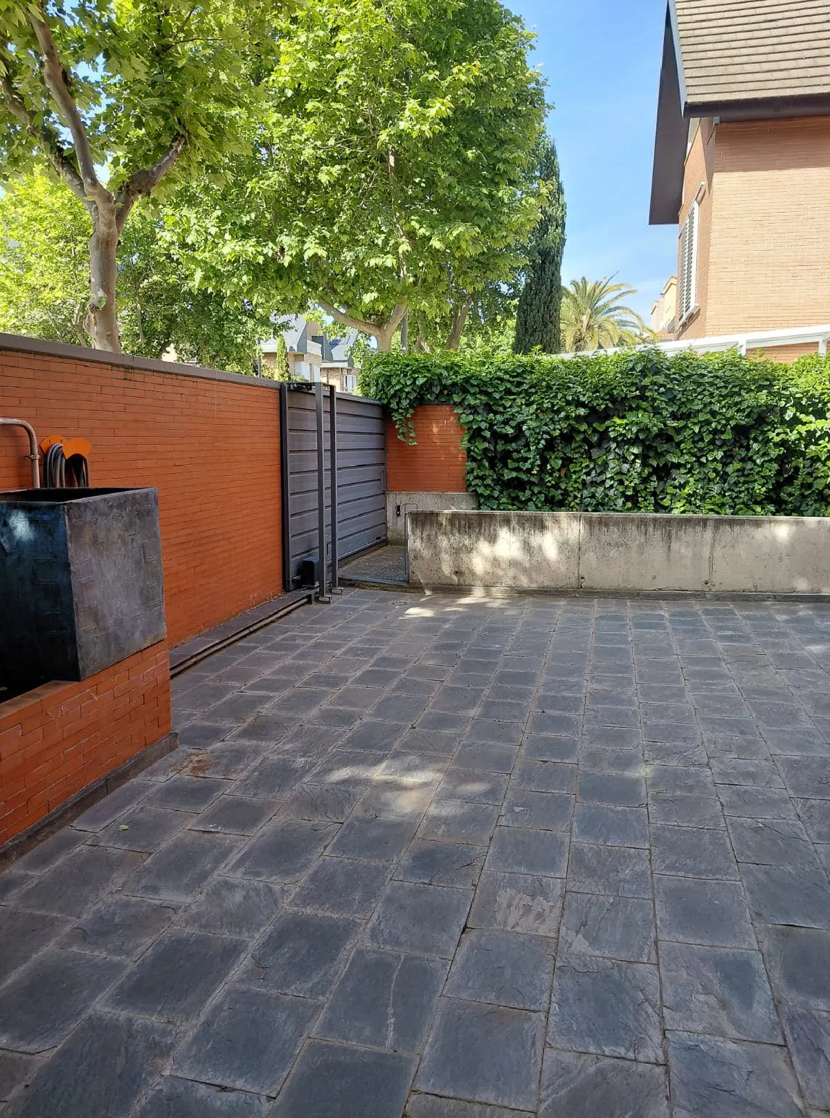 A stone-paved courtyard with a brick wall, a gray gate, and green foliage. A stone planter sits on the left.